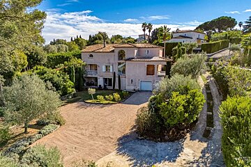 A large Mediterranean-style villa set amidst abundant greenery and trees under a clear blue sky, with a spacious gravel driveway.