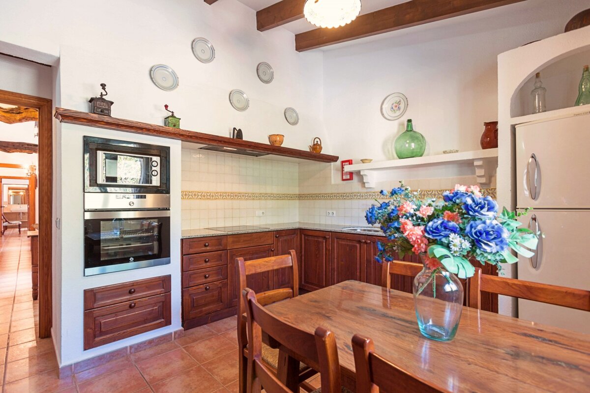 A traditional kitchen with dark wooden cabinets, a terracotta tiled floor, and a dining table with a vibrant floral centerpiece. White walls display decorative plates.