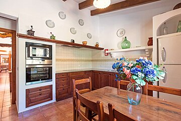 A traditional kitchen with dark wooden cabinets, a terracotta tiled floor, and a dining table with a vibrant floral centerpiece. White walls display decorative plates.