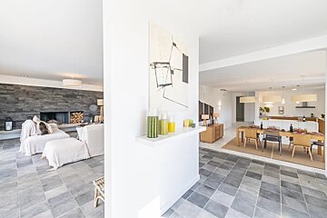 Modern open-plan living space with a stone fireplace, white sofas, dining area, and kitchen. Features gray tiled floor, white walls, and modern art.