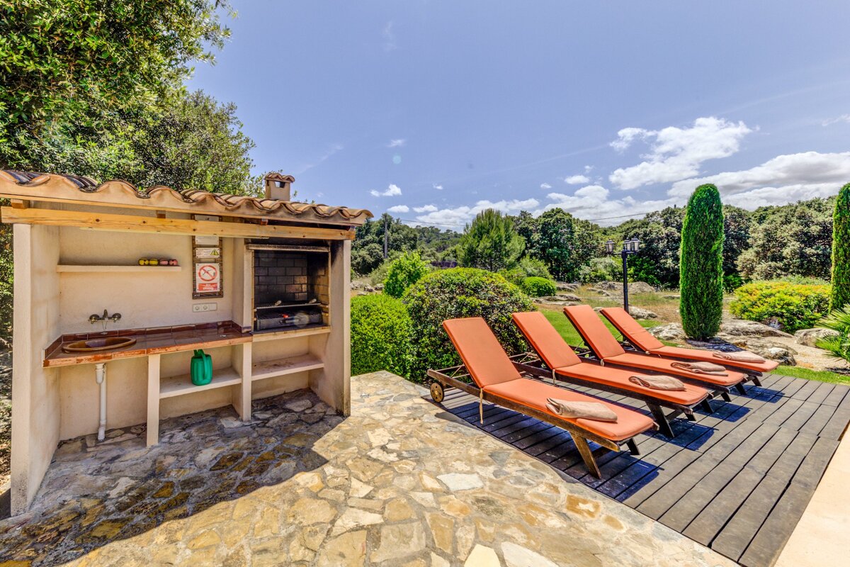 Sunny outdoor living space with a BBQ, stone patio, and four orange lounge chairs on a deck, set against a lush green garden under a blue sky.