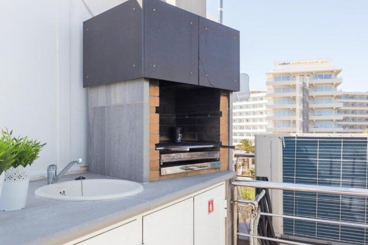 An outdoor balcony kitchen featuring a built-in stone barbecue grill, a sink, and cabinets, with city buildings in the background.