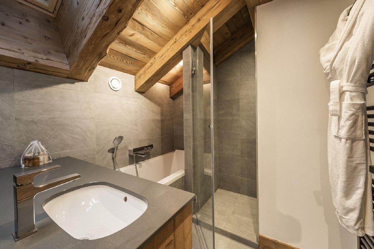 A chalet-style bathroom with exposed wooden beams, grey tiles, a bathtub, glass shower, sink vanity, and a white robe hanging.