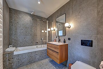 Stylish bathroom featuring grey speckled tiles, a white bathtub, floating wood vanity, and contemporary mirror with spherical lights.