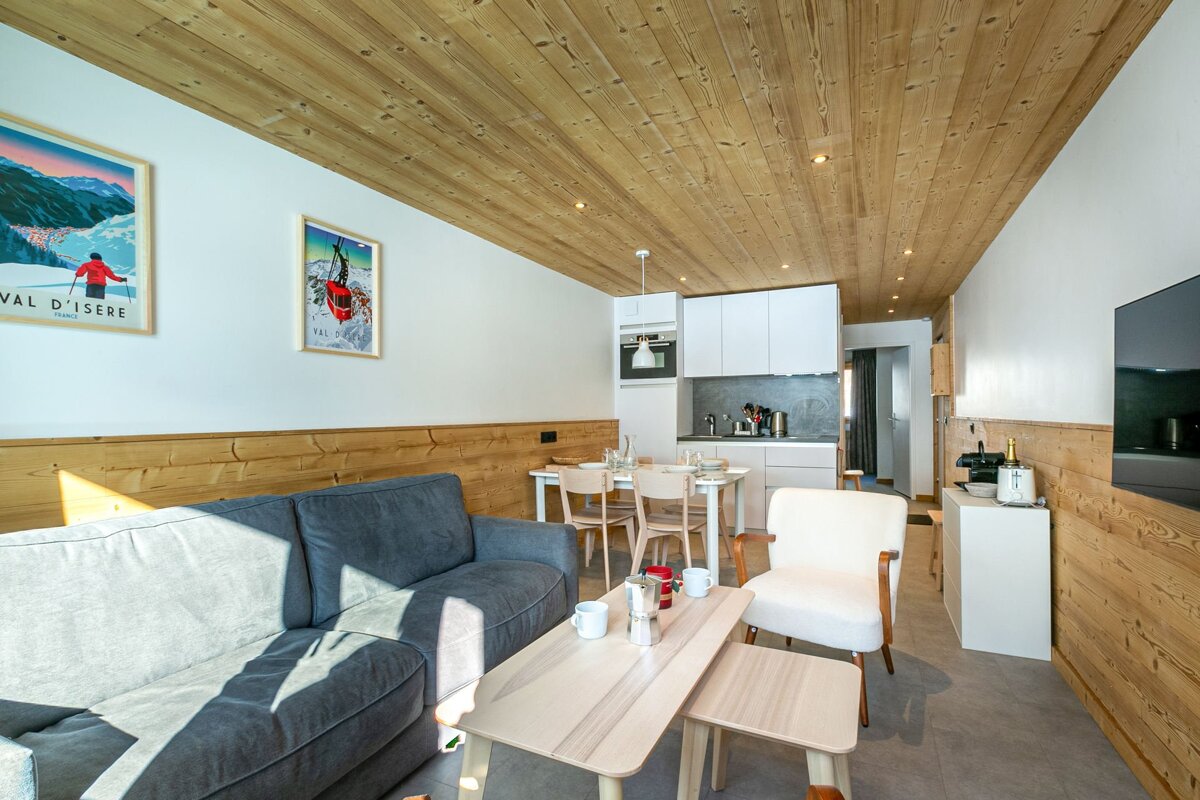 A modern open-plan living and kitchen area featuring wood-paneled ceilings, white walls with ski posters, and a cozy grey sofa. Rustic-chic interior.