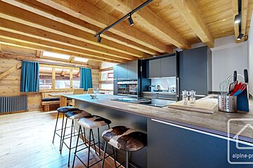 A modern kitchen in a rustic wooden chalet, featuring dark cabinetry, a large island with cowhide stools, exposed wooden beams, and a window with blue curtains.