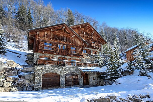 Rustic wooden chalets with stone bases sit on a snowy mountain slope, surrounded by pine trees under a clear blue sky.