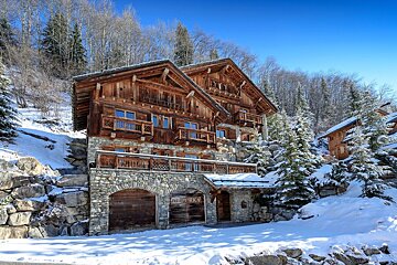 Rustic wooden chalets with stone bases sit on a snowy mountain slope, surrounded by pine trees under a clear blue sky.