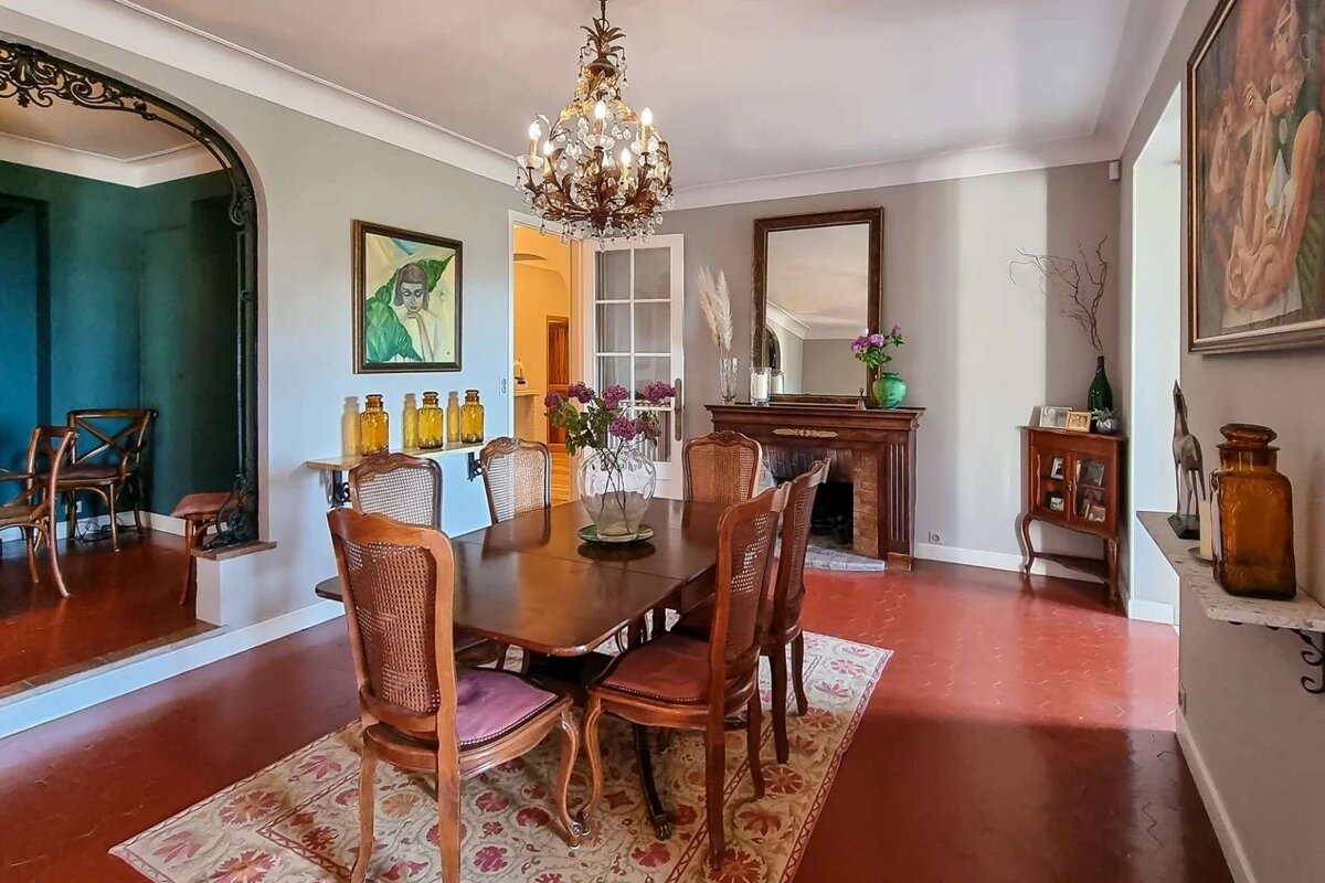 A warm, traditional dining room with a long wooden table, elegant chandelier, fireplace, and reddish floor. Walls are grey, decorated with art.
