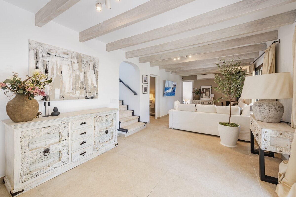A bright, open living area with light wood beams, a rustic white sideboard, and abstract art. Neutral tones and a white sofa define the serene space.