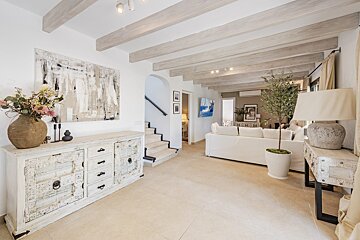 A bright, open living area with light wood beams, a rustic white sideboard, and abstract art. Neutral tones and a white sofa define the serene space.