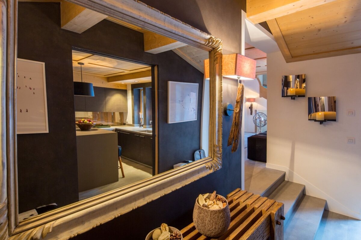 A large ornate mirror reflects a modern kitchen. Adjacent, a cozy hallway features stairs, sconces, a wooden bench with potpourri.