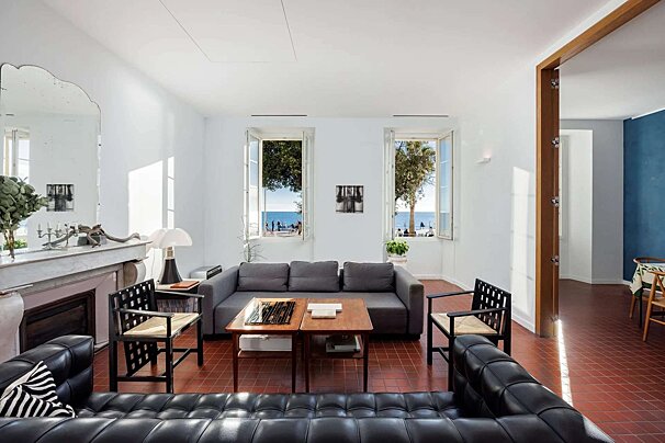 Bright, modern living room with a sea view from open windows. Features black and grey sofas, unique chairs, and terracotta floor tiles.