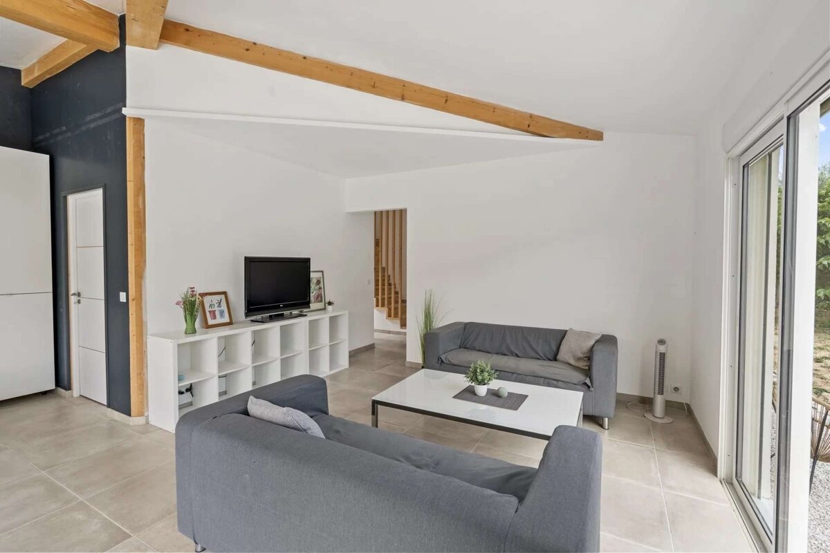 A bright, modern living room featuring white walls, exposed wood beams, two gray sofas, a coffee table, a TV, and a large sliding glass door.