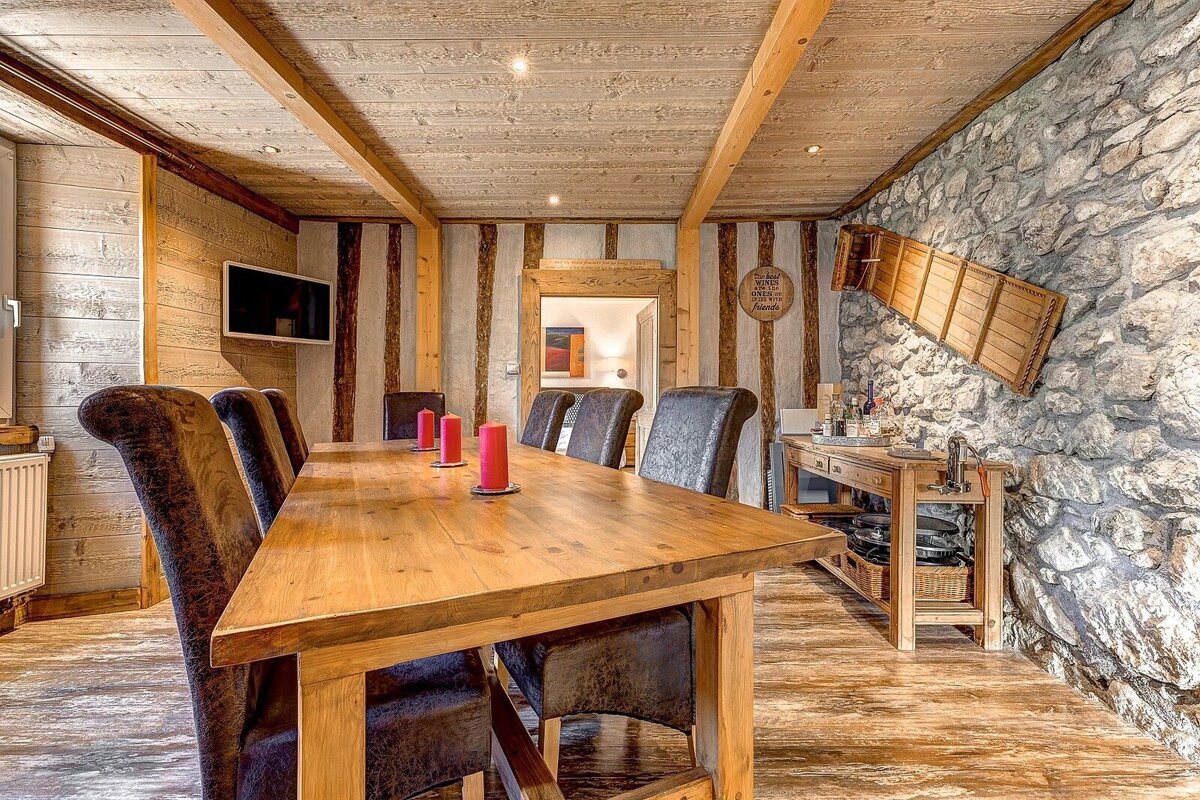 A cozy, rustic dining room features a large wooden table, leather chairs, wood-paneled walls/ceiling, and a prominent stone wall with ski decor.