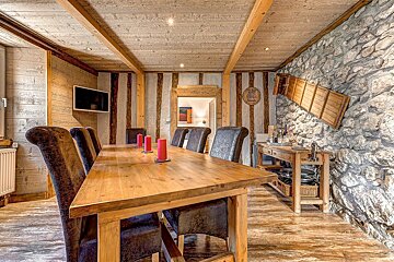 A cozy, rustic dining room features a large wooden table, leather chairs, wood-paneled walls/ceiling, and a prominent stone wall with ski decor.