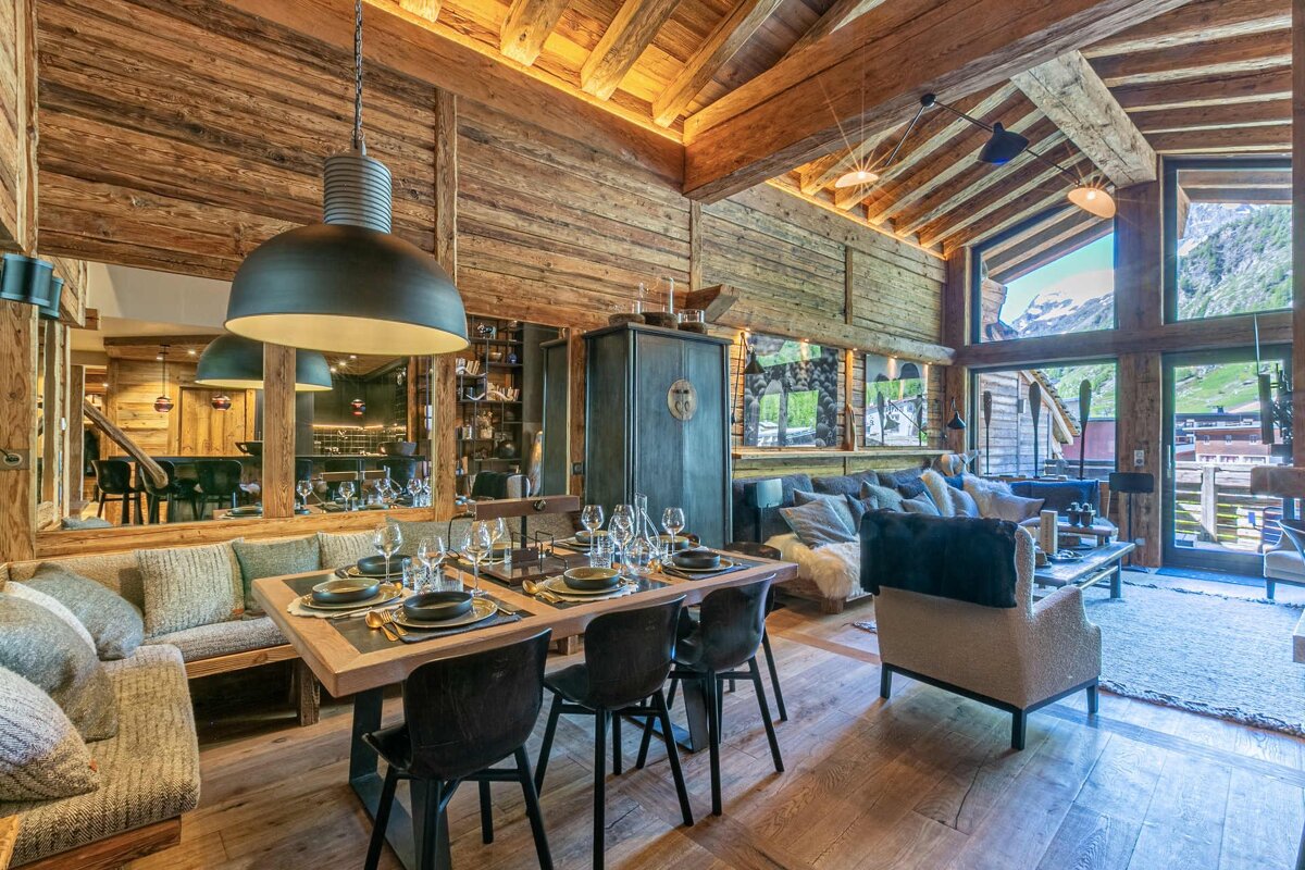 Luxurious, rustic wooden chalet interior featuring a set dining table, cozy living room, and large windows with stunning mountain views.