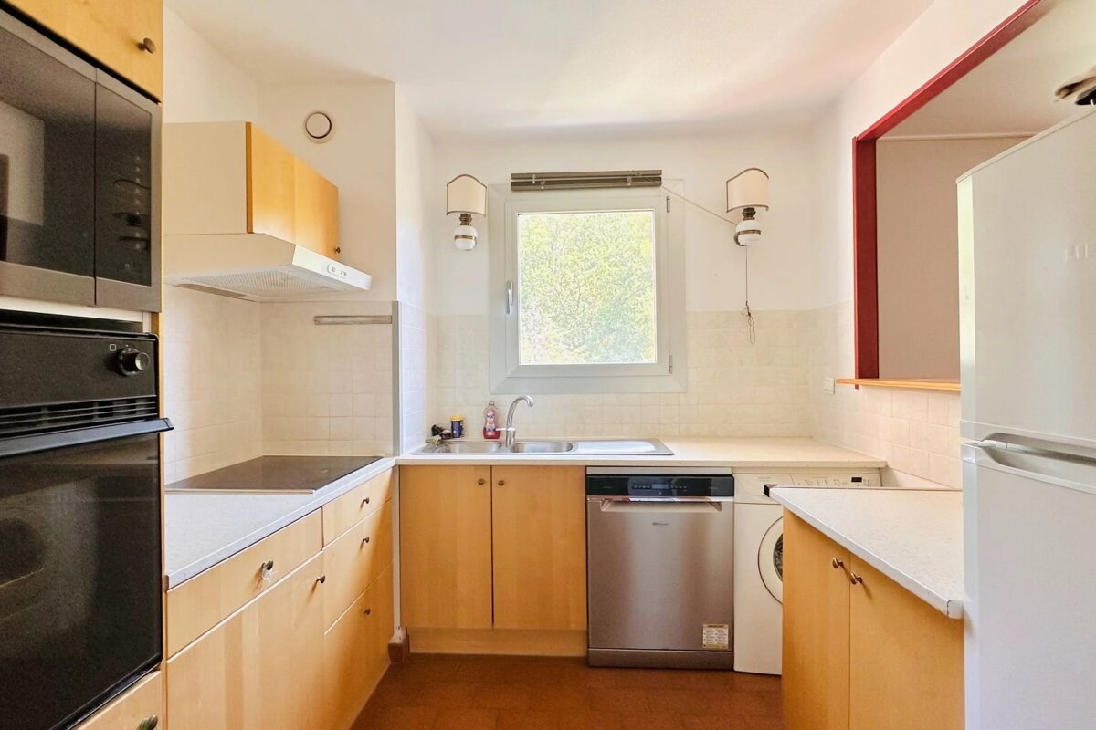 A small kitchen with light wood cabinets, a window over the sink, a dishwasher, washing machine, and refrigerator.