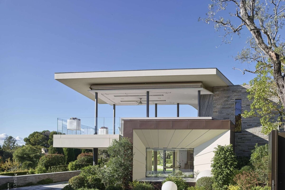 A modern multi-level house with flat roofs, glass railings, and expansive windows, surrounded by green foliage under a clear blue sky.