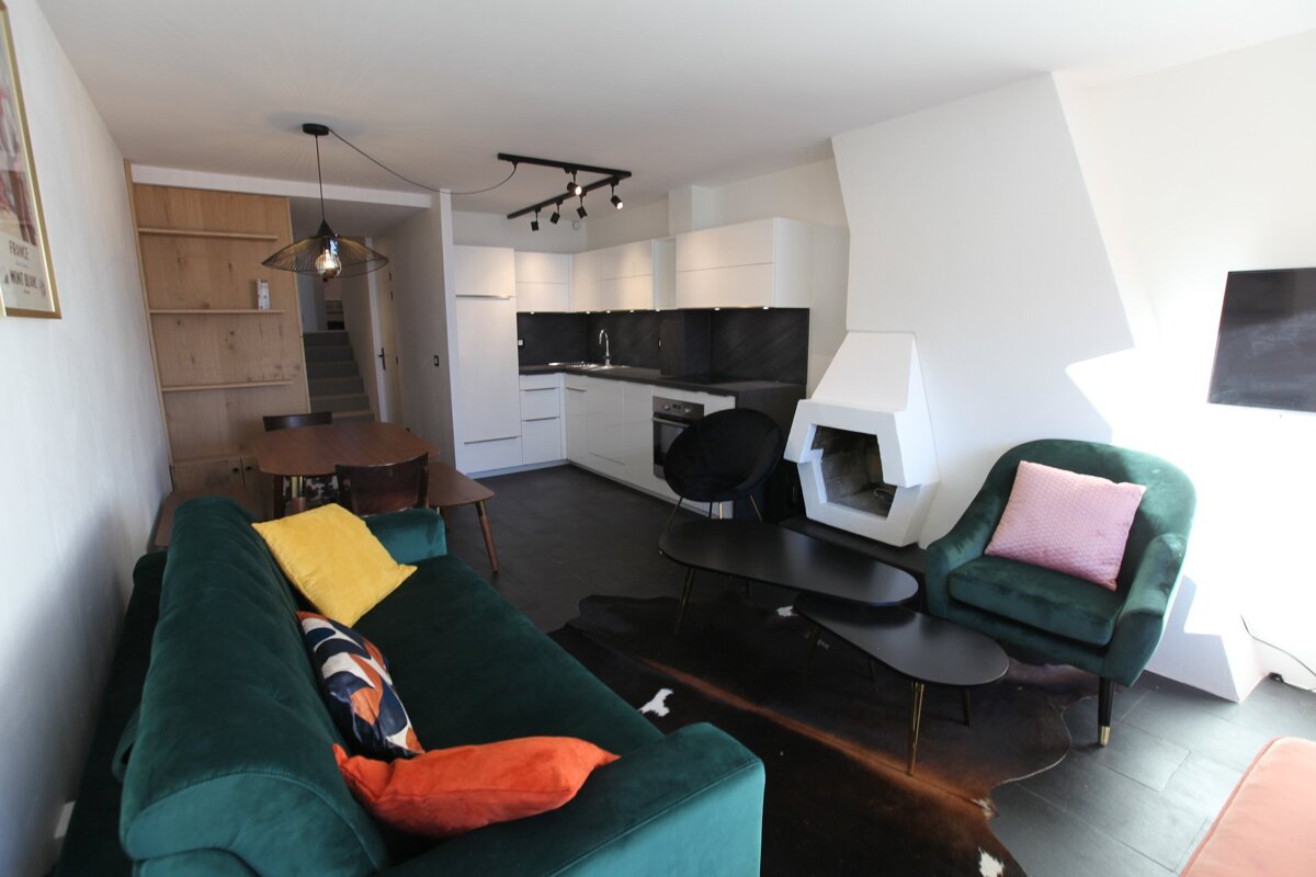 A modern open-plan living space with a white kitchen, dark green velvet sofa and armchair, unique white fireplace, and a dining area.
