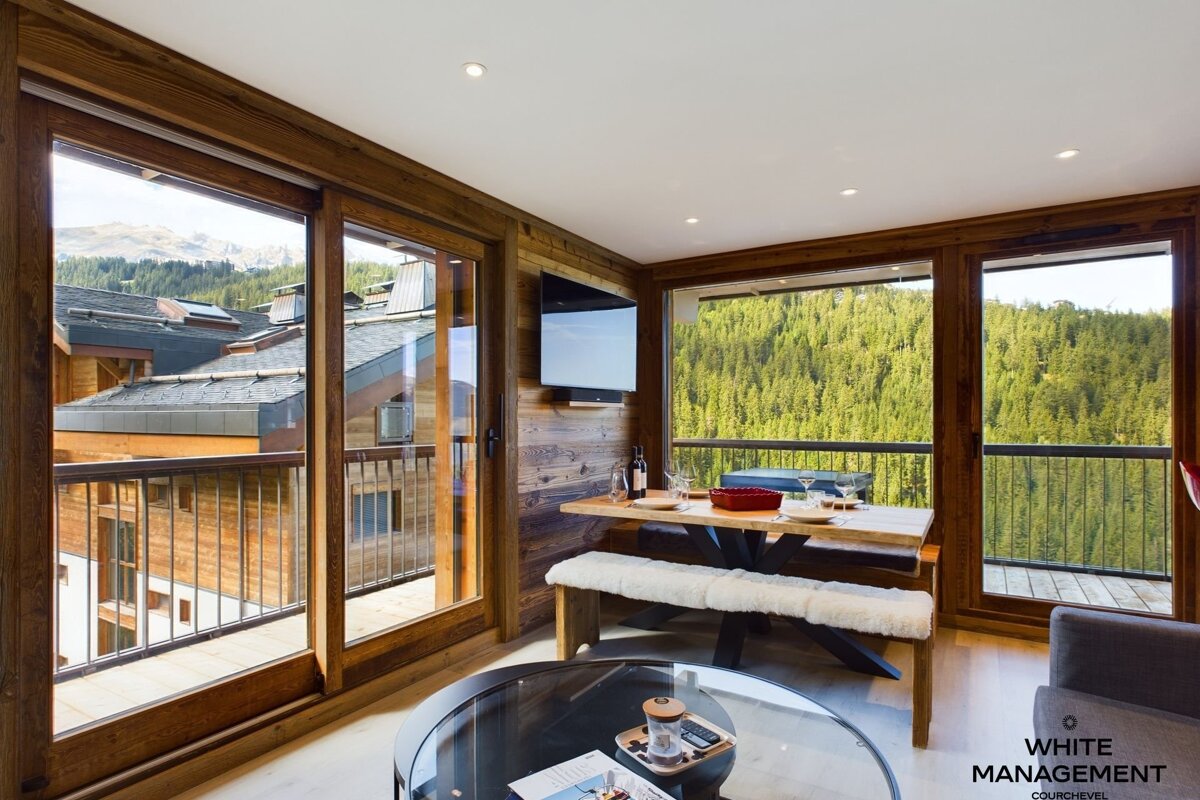 Rustic modern living/dining area with wood paneling, large windows, and balconies offering stunning mountain and forest views. Features a dining table, TV, and coffee table.
