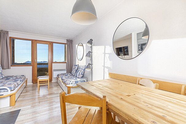Bright living/dining room with a wooden table, two sofa beds, and large windows offering a mountain view. Mirrors adorn the white walls.