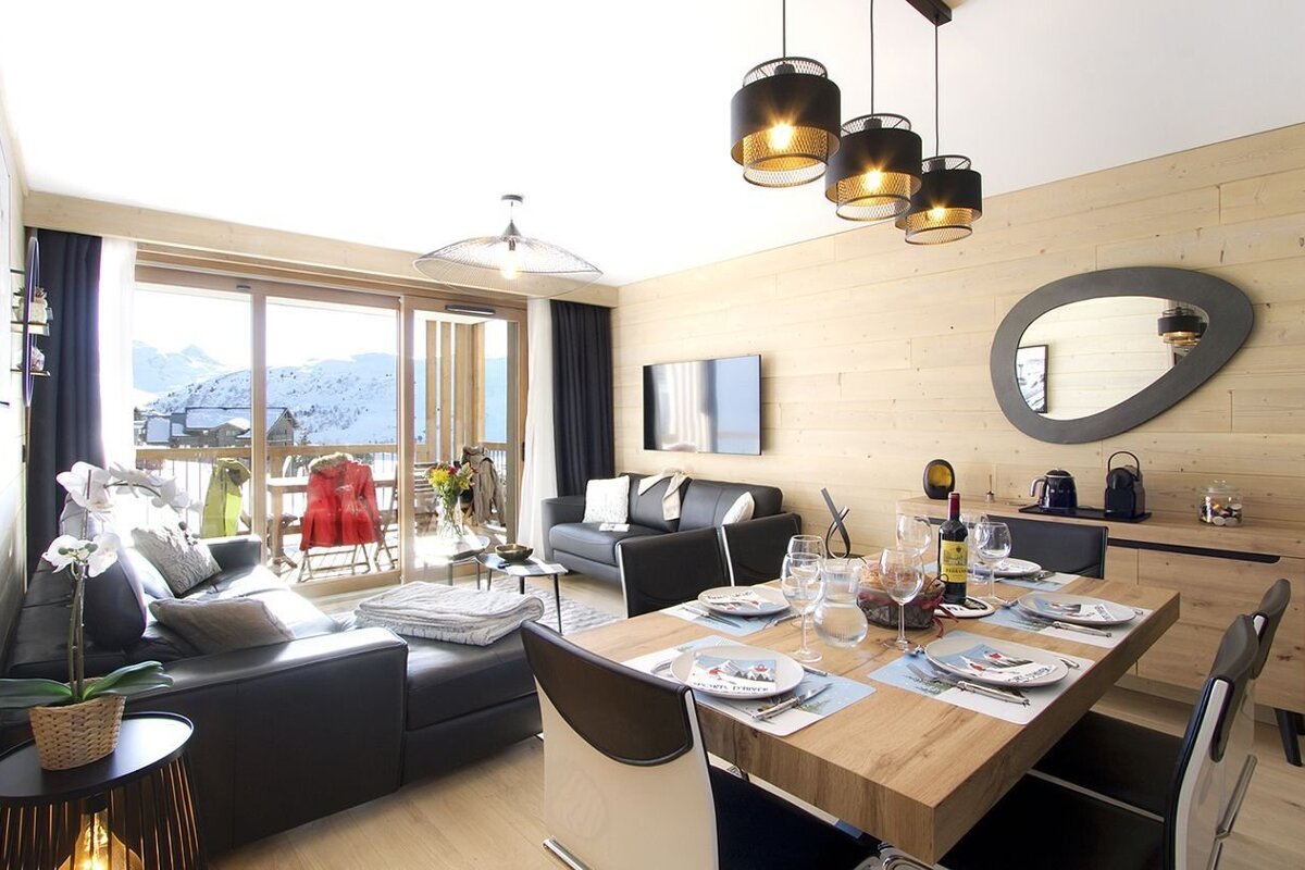 A modern living/dining room features dark sofas, a set dining table, and a balcony with snow-covered mountain views. Light wood walls and contemporary lighting add warmth.