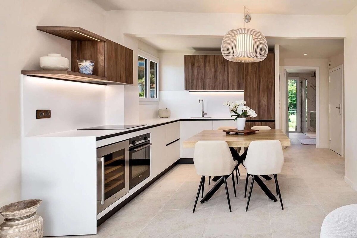 A sleek, modern kitchen featuring white and wood cabinetry, integrated appliances, and a light wooden dining table with cream chairs under a stylish pendant lamp.