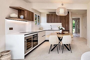 A sleek, modern kitchen featuring white and wood cabinetry, integrated appliances, and a light wooden dining table with cream chairs under a stylish pendant lamp.