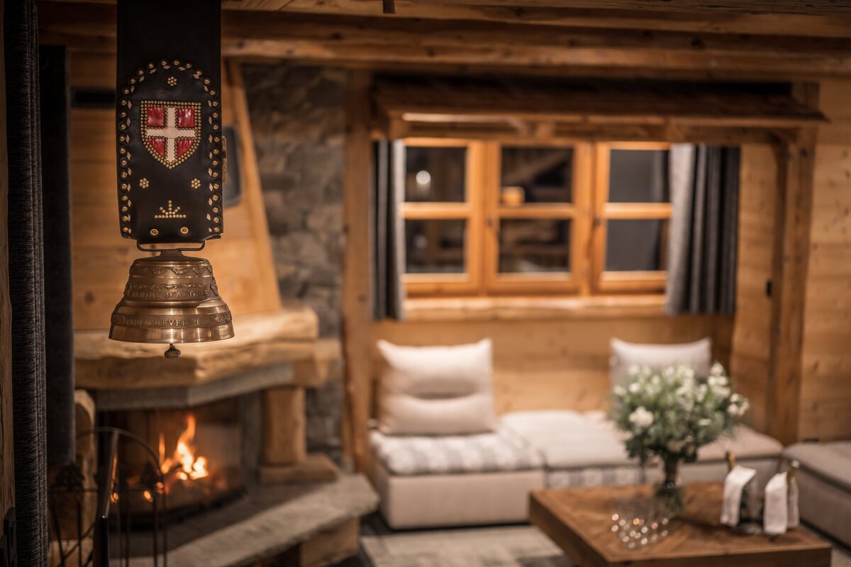 Cozy alpine chalet interior with a burning fireplace, wooden furniture, and a decorative cowbell hanging in the foreground.