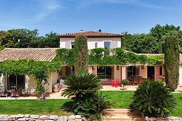 A Mediterranean villa with a terracotta roof, lush green vines, sago palms, and a manicured lawn under a clear blue sky.
