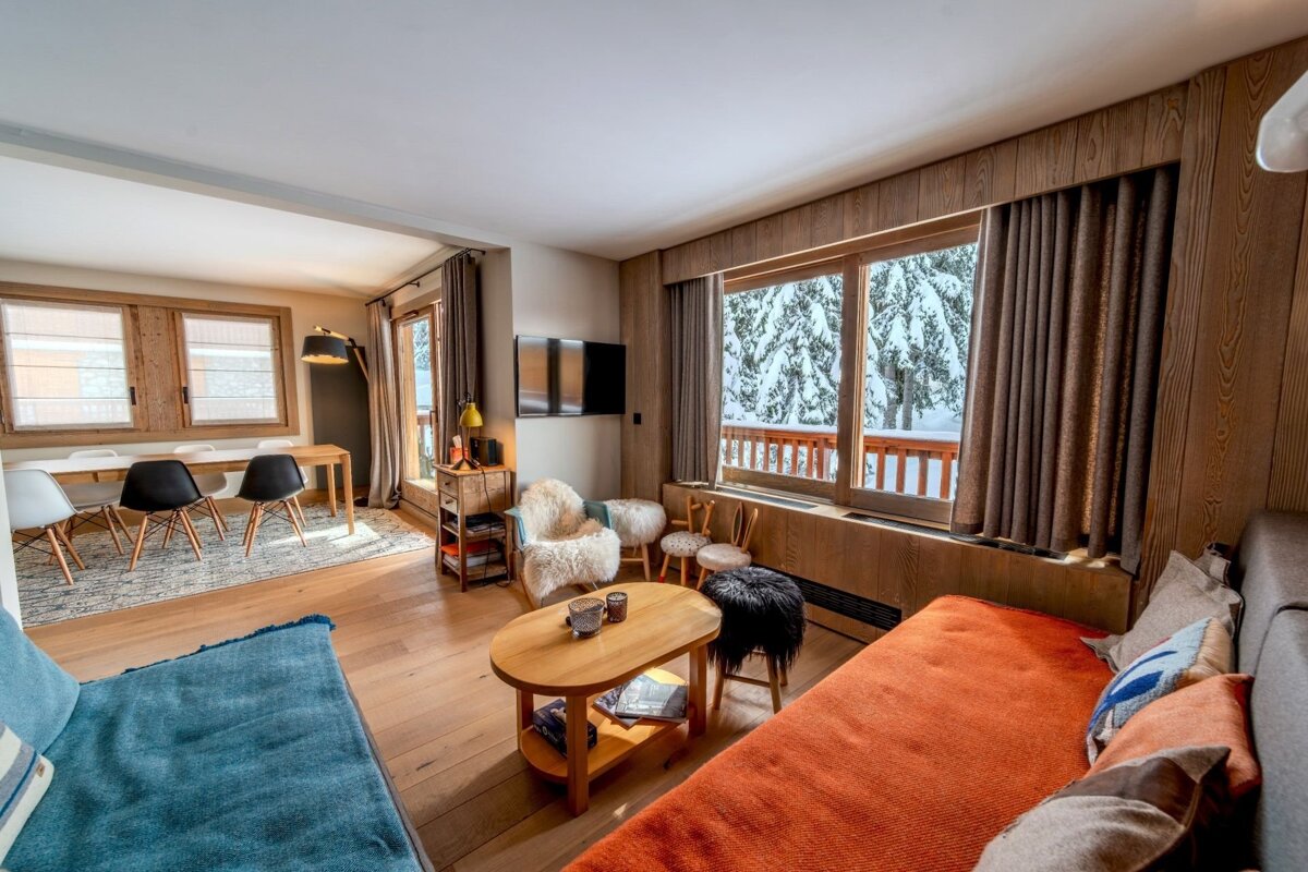 Cozy alpine cabin interior with wood paneling, an orange sofa, and a large window overlooking a snowy winter forest.