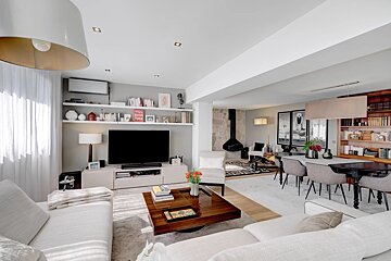 Bright, modern open-plan living and dining room with white sofas, a large TV, bookshelves, wooden coffee table, and a black hanging fireplace.