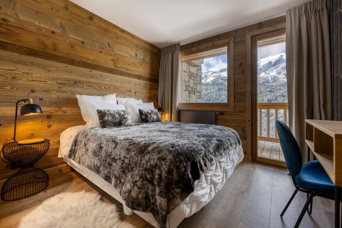 Cozy chalet bedroom with wooden walls, a large bed with a faux fur throw, and a stunning view of snowy mountains from the window and balcony.