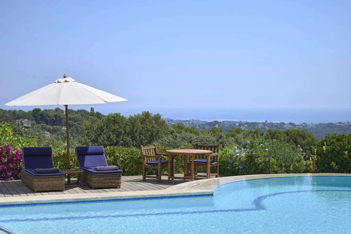 Luxurious poolside terrace with blue lounge chairs, a dining set, and umbrella, offering stunning views of a green landscape and the distant sea on a sunny day.