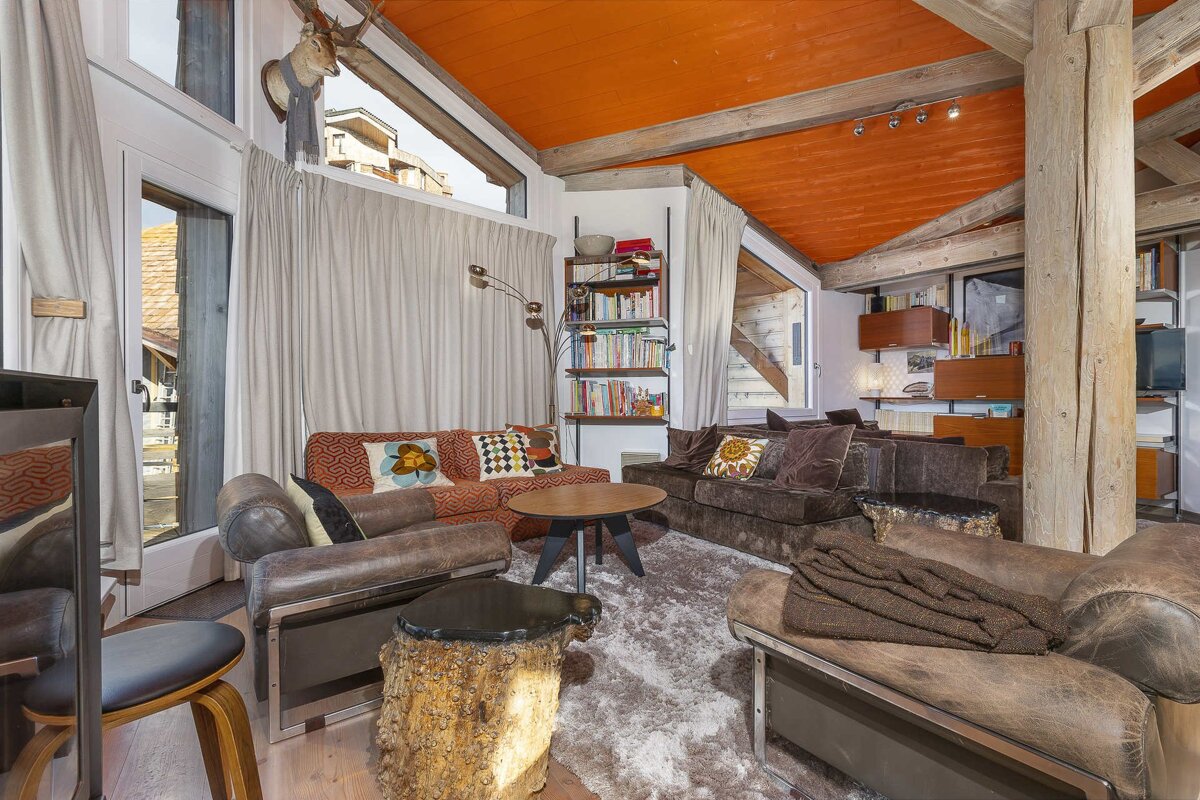A modern rustic living room with a vibrant orange ceiling, exposed wooden beams, plush sofas, a deer head, and a unique tree stump table.