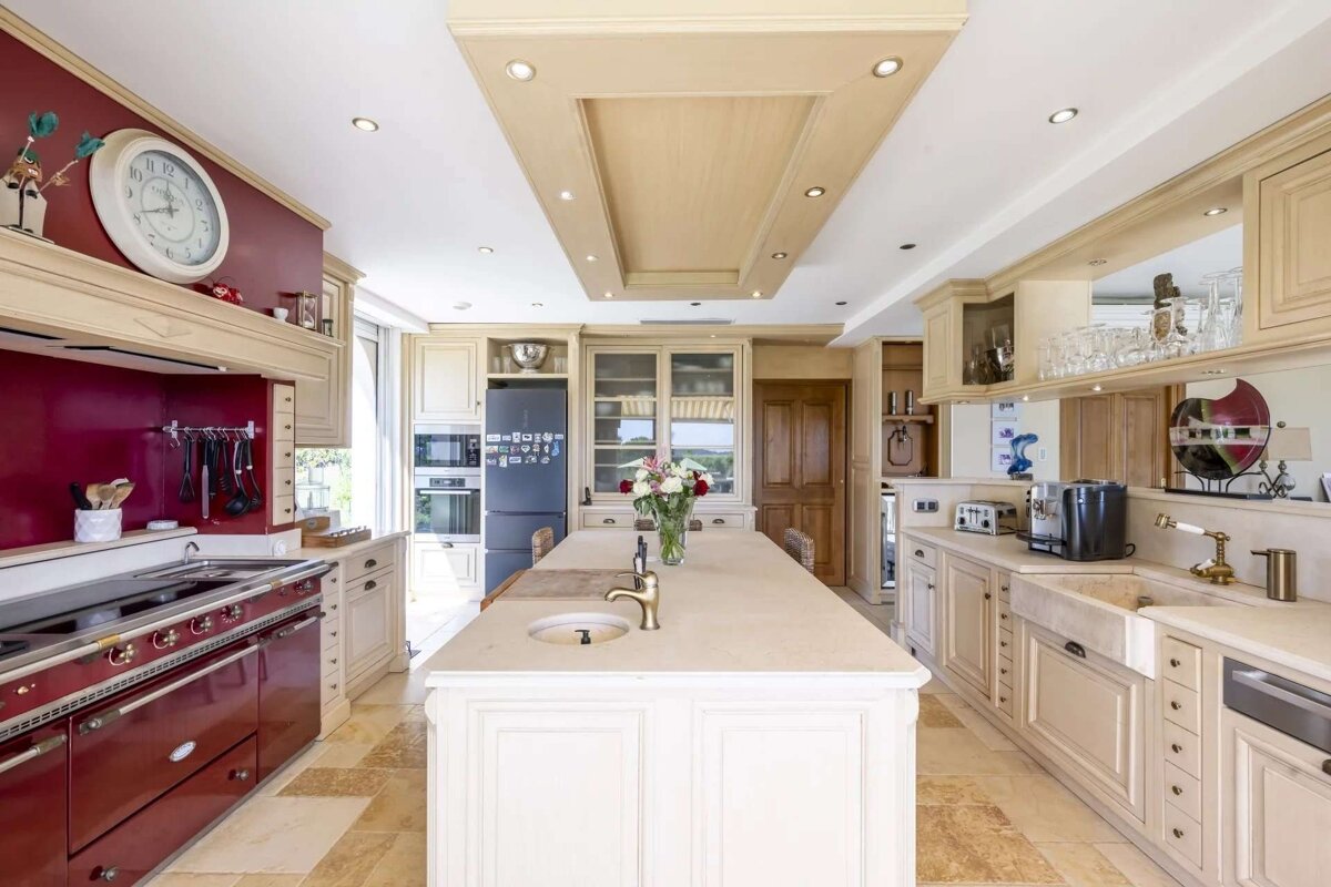 A bright, spacious kitchen with cream cabinets, a large central island, and a prominent red stove. Features modern appliances and tiled flooring.