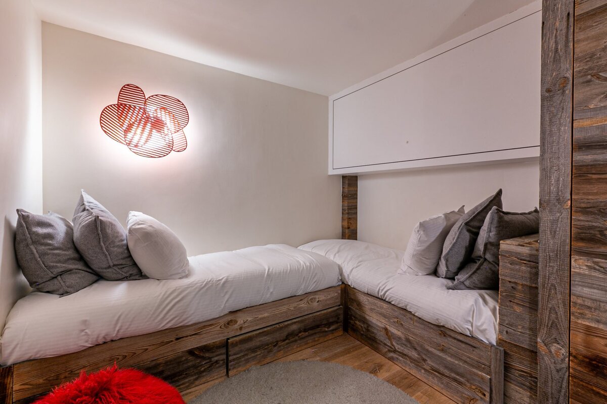 Cozy room with two rustic wooden beds, white bedding, grey pillows, and a vibrant red artistic wall light illuminating the neutral walls.
