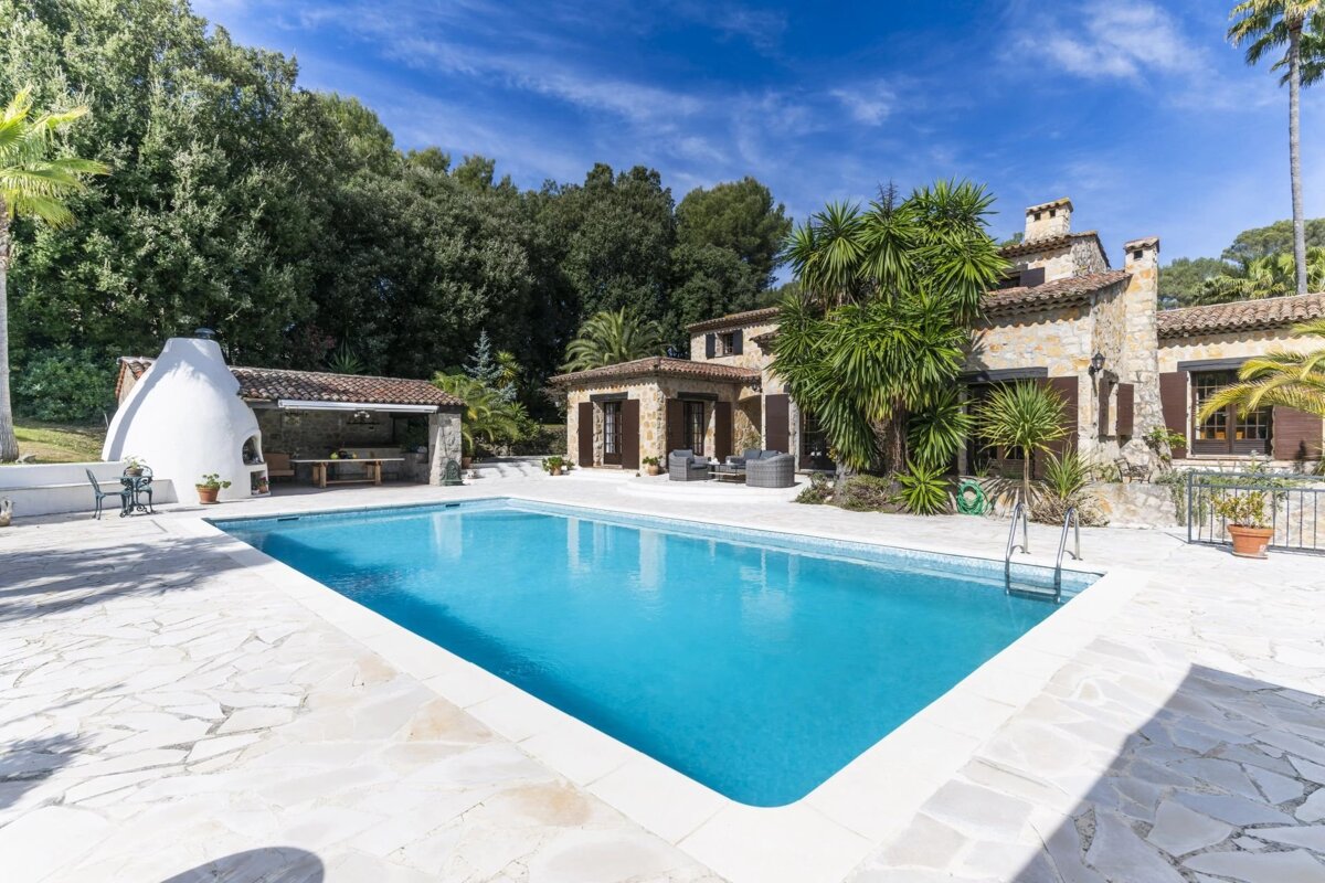 A beautiful stone villa with a large swimming pool, outdoor kitchen, and lush green landscaping under a bright blue sky.