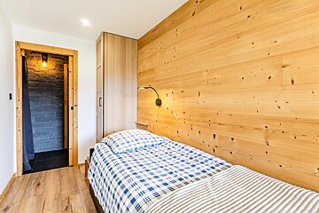Cozy rustic bedroom with a wood-paneled wall, a single bed in blue plaid, and a doorway leading to a stone bathroom.