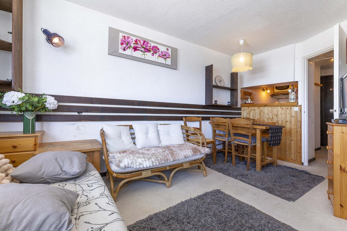 A cozy, compact living space featuring a rattan sofa, wooden dining set, bed, and a small kitchenette with wooden paneling.