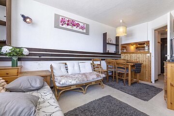 A cozy, compact living space featuring a rattan sofa, wooden dining set, bed, and a small kitchenette with wooden paneling.
