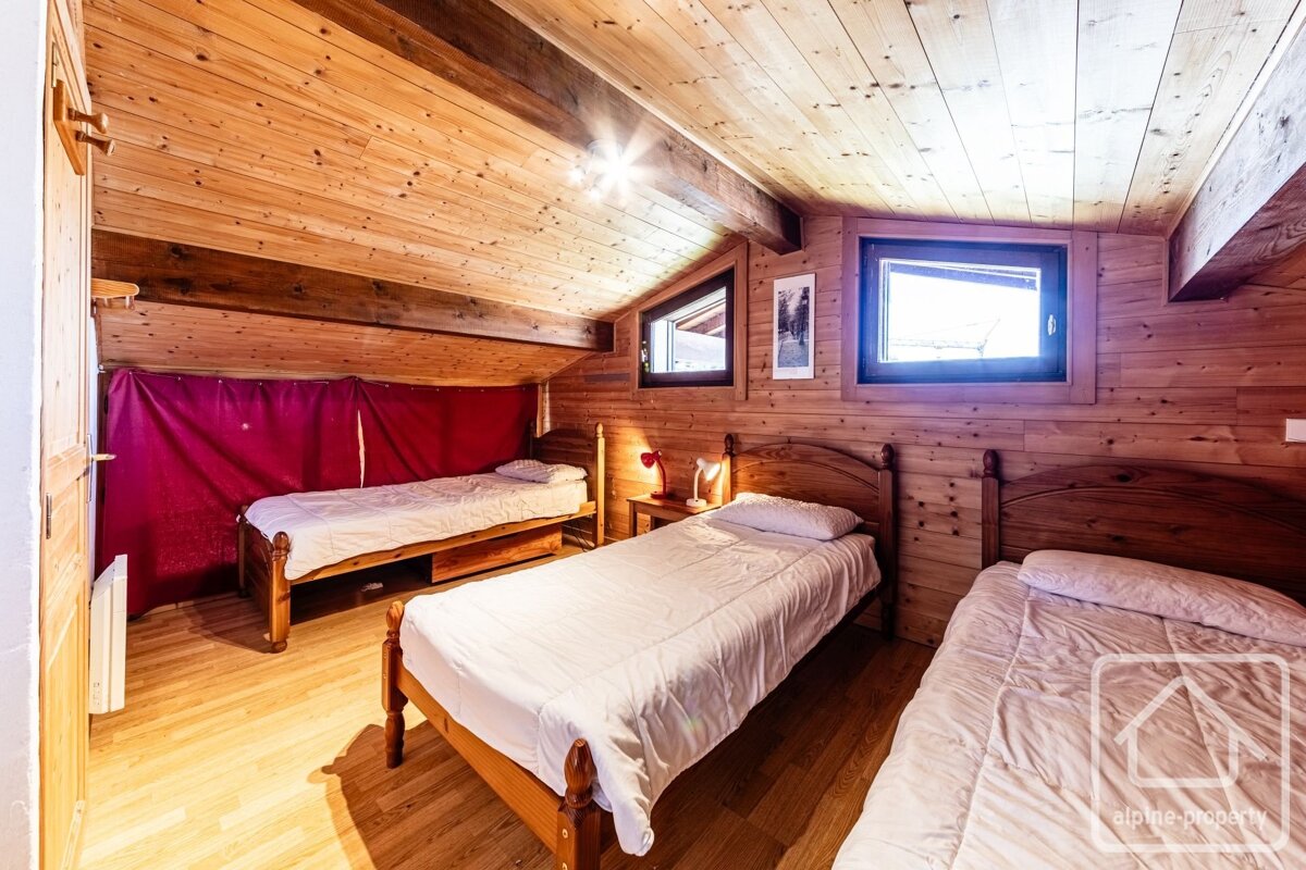 Bright, rustic attic bedroom with wooden walls and ceiling, featuring three single beds, two windows, and a red curtain.