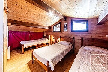 Bright, rustic attic bedroom with wooden walls and ceiling, featuring three single beds, two windows, and a red curtain.