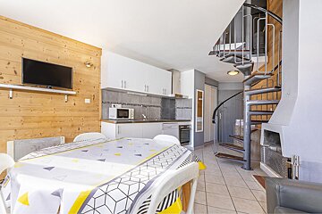 An open-plan living space featuring a modern kitchen, dining table, wood-paneled wall with a TV, and a grey metal spiral staircase on tiled floors.