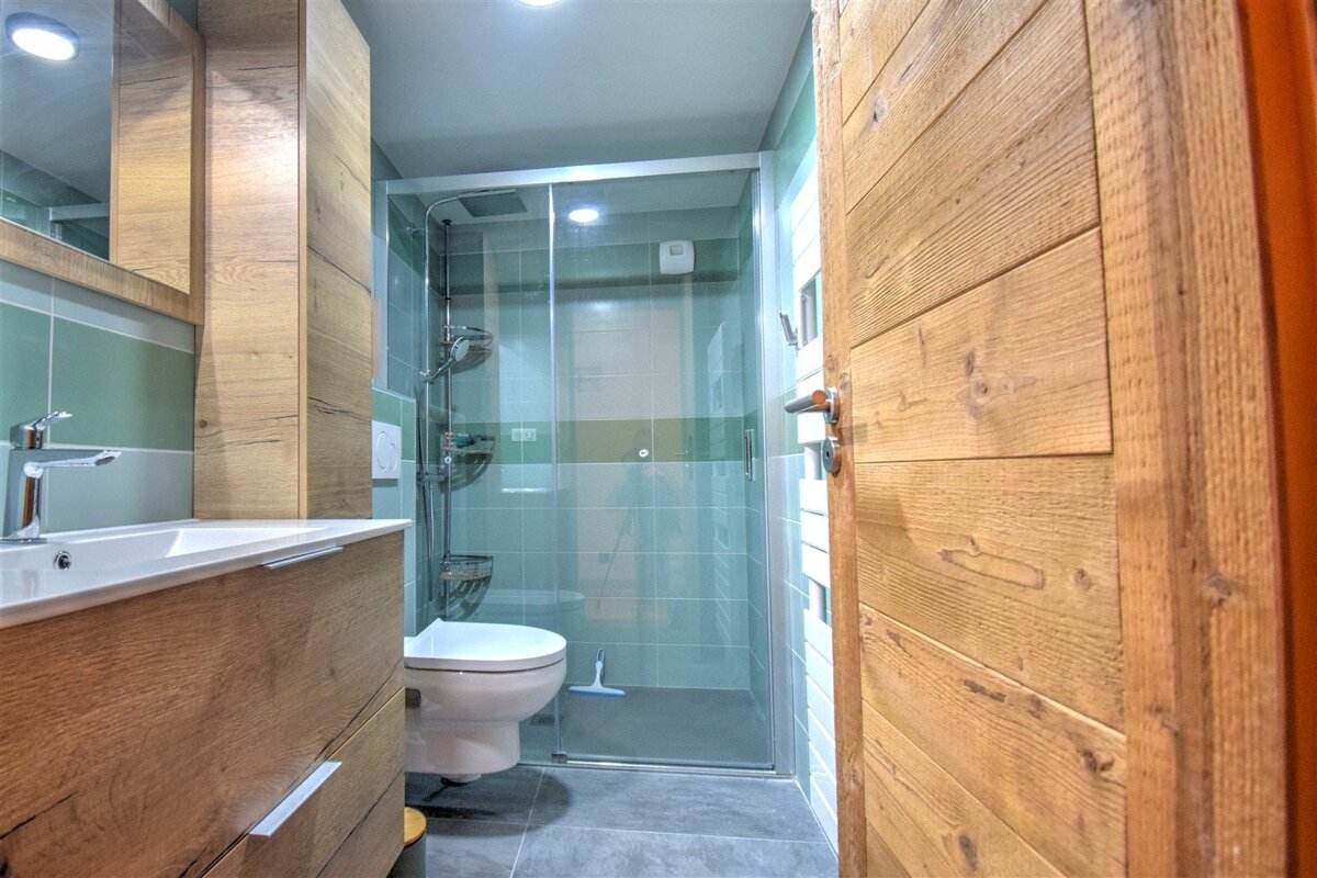 A modern bathroom featuring a wooden vanity, sink, and cabinets, green and white tiled walls, a glass shower enclosure, toilet, and a light wooden door.