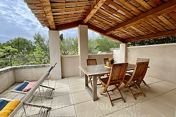 A serene, covered outdoor patio features a wooden dining set, two lounge chairs, and a lush green landscape visible under a tiled roof.