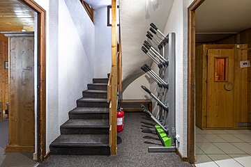 A ski lodge boot room with a carpeted staircase, wooden railing, and a multi-slot ski boot dryer. A fire extinguisher and a sauna door are also visible.