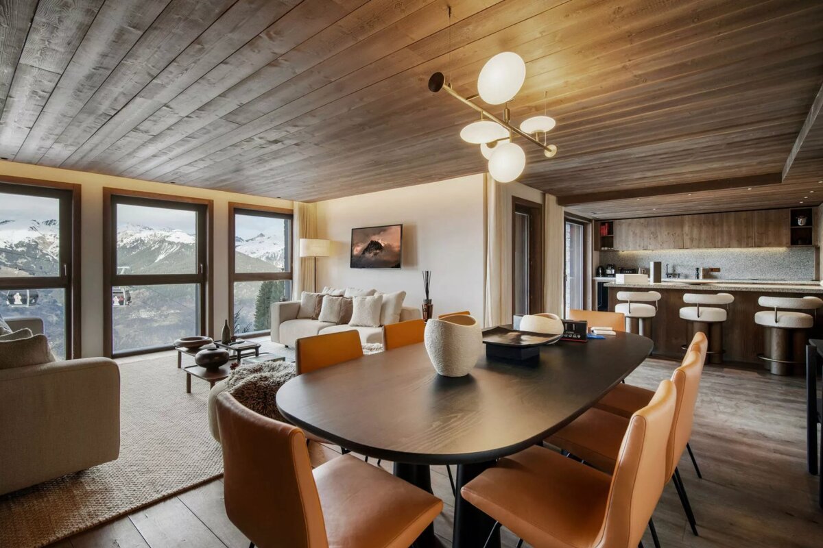 Modern open-plan living, dining, and kitchen area with panoramic snowy mountain views through large windows and a stylish wooden ceiling.