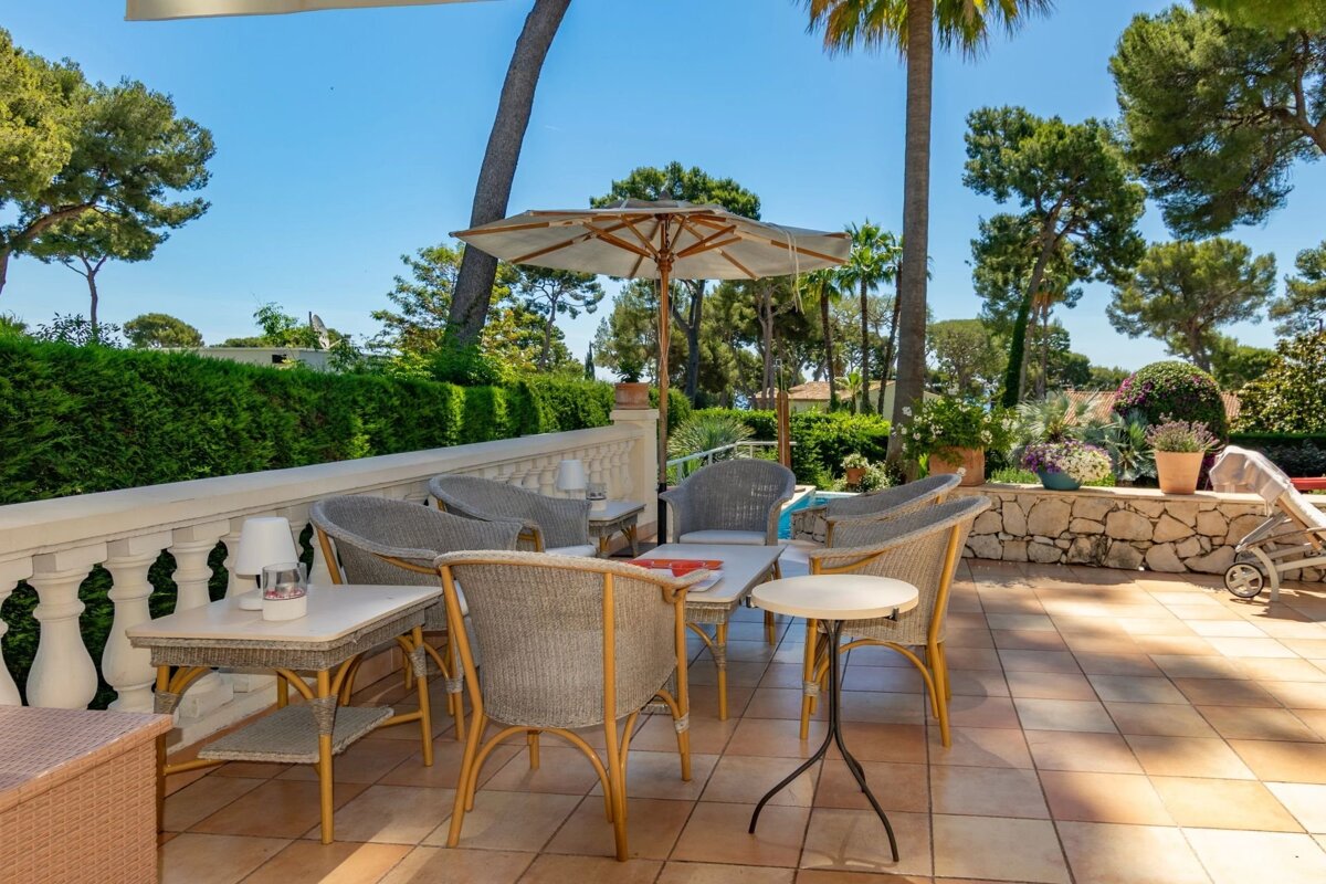Sunny tiled terrace with wicker armchairs, tables, and an umbrella, surrounded by lush greenery and palm trees under a bright blue sky.